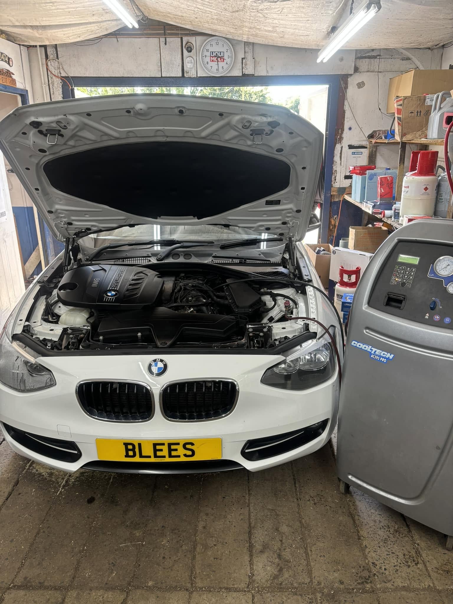 A white BMW with its bonnet up beside the air conditioning machine