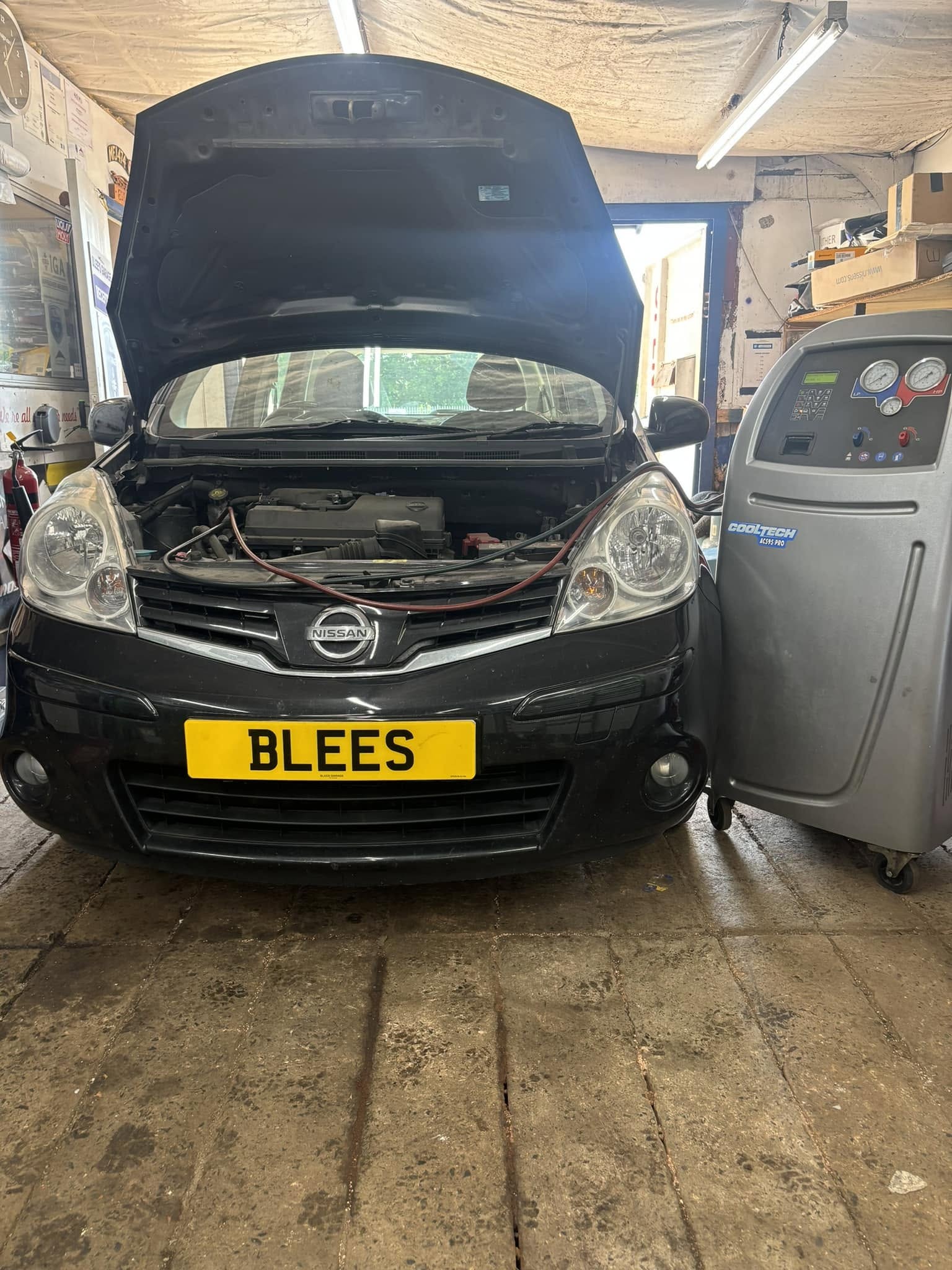 A black Nissan in the workshop bay with its bonnet up, the air conditioning machine beside it
